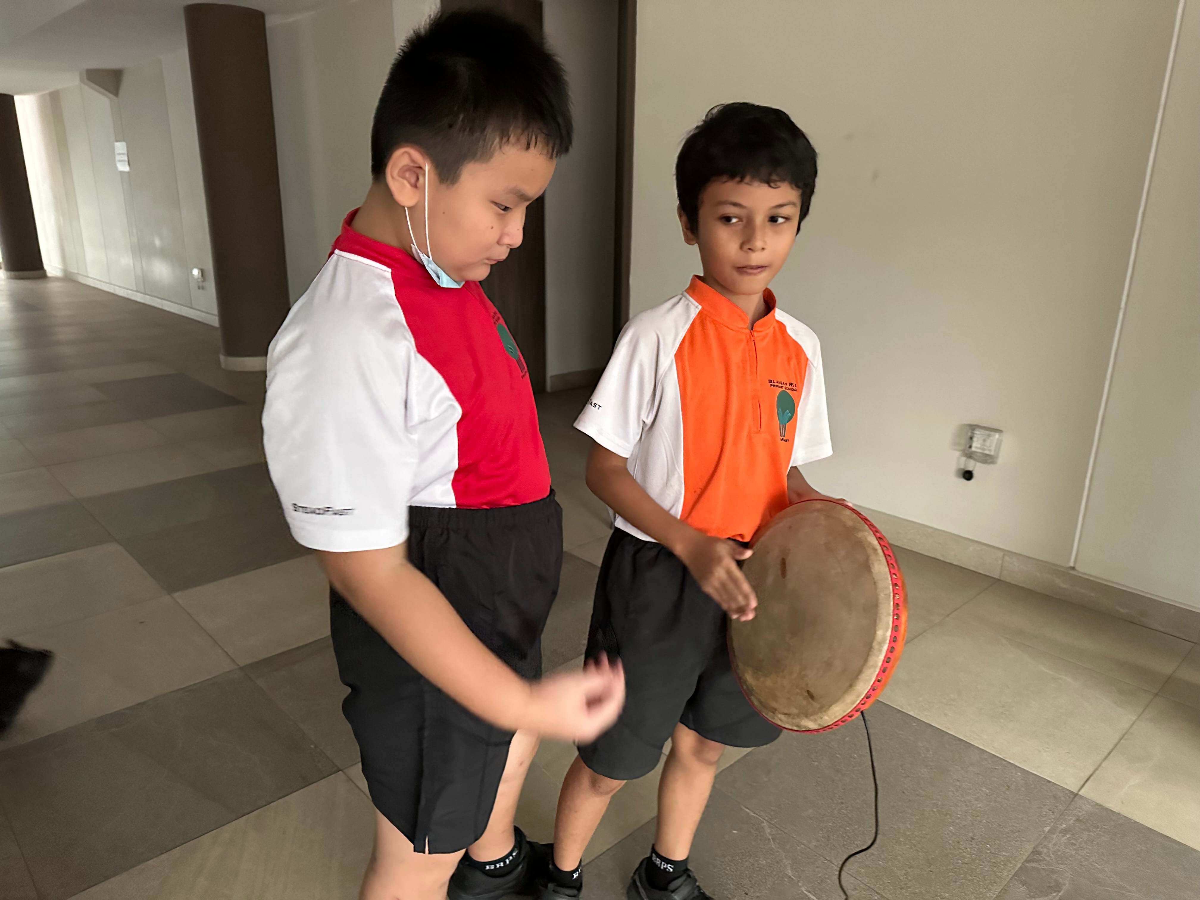 Two boys in school uniforms stand in a corridor, one holding a round hand drum while the other appears to be playing it.